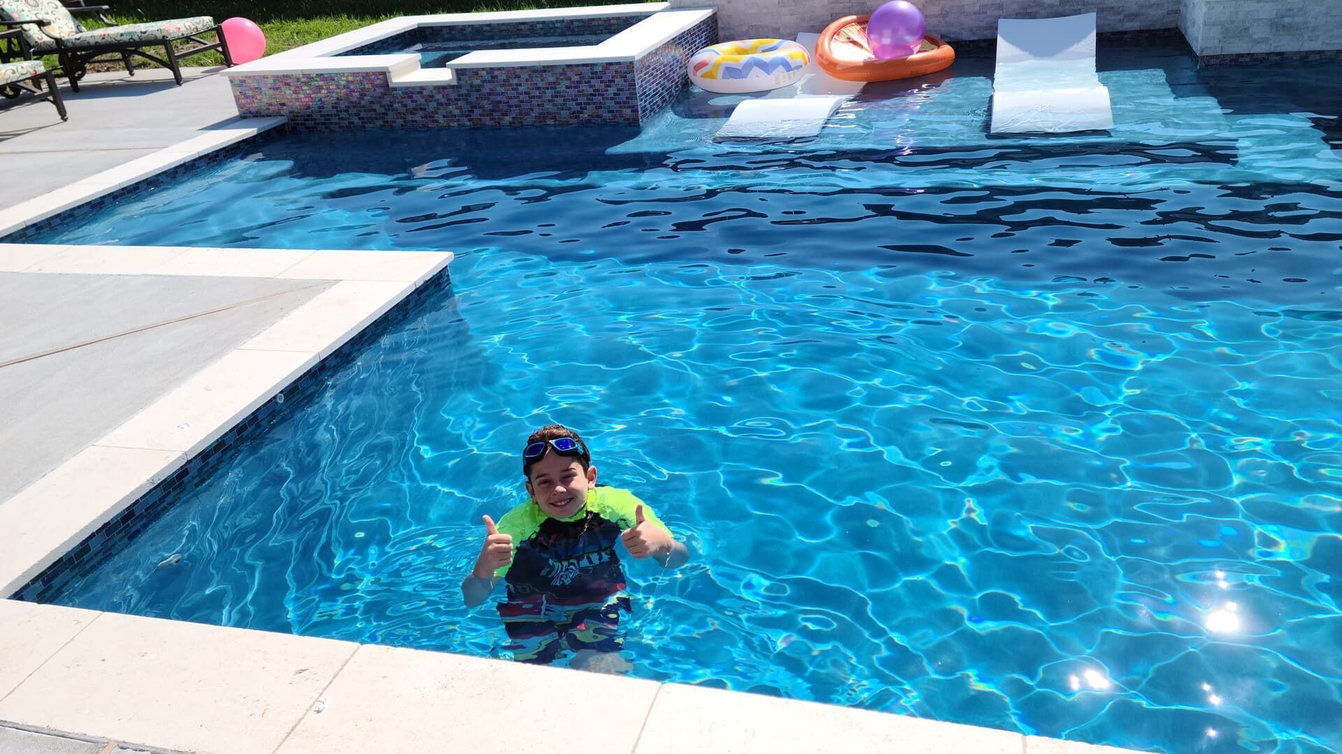 Child in a blue pool wearing a neon vest, with pool floats and a slide in the background.