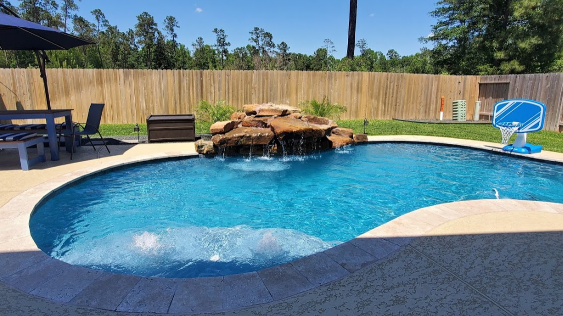 Backyard swimming pool with waterfall rock feature, blue water, patio chairs, and a wooden fence