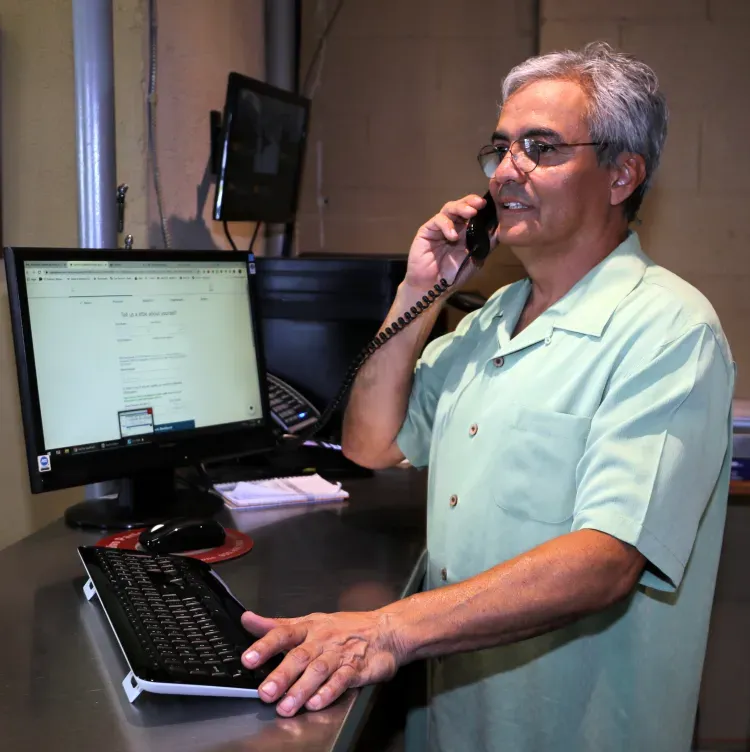 Man in a light green shirt talking on a desk phone beside a computer monitor and keyboard in an office