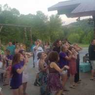 People dancing outdoors at a lively gathering, with a speaker and greenery in the background