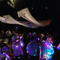 People at a nighttime outdoor event dancing under colorful lights and hanging white fabric canopies