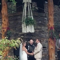 Bride and groom exchange vows under a rustic wooden arch with an officiant at an outdoor ceremony