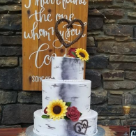 Three-tier white marbled cake with sunflowers and roses on a rustic table, in front of a wooden sign.