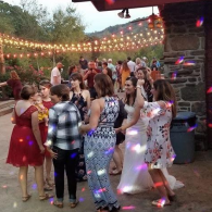 People dancing at an outdoor party under string lights, with a stone wall and trees in the background.