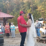 Couple in red shirt and white dress sharing a dance outdoors at a rustic gathering.
