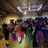 People dancing at a party in a banquet hall with colorful lights and a DJ booth in the background