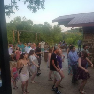 Group dancing outdoors near a rustic building at dusk, with trees and hills in the background