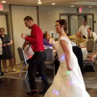 Bride in white dress dancing at a reception with a man in a red shirt, seated guests in the background