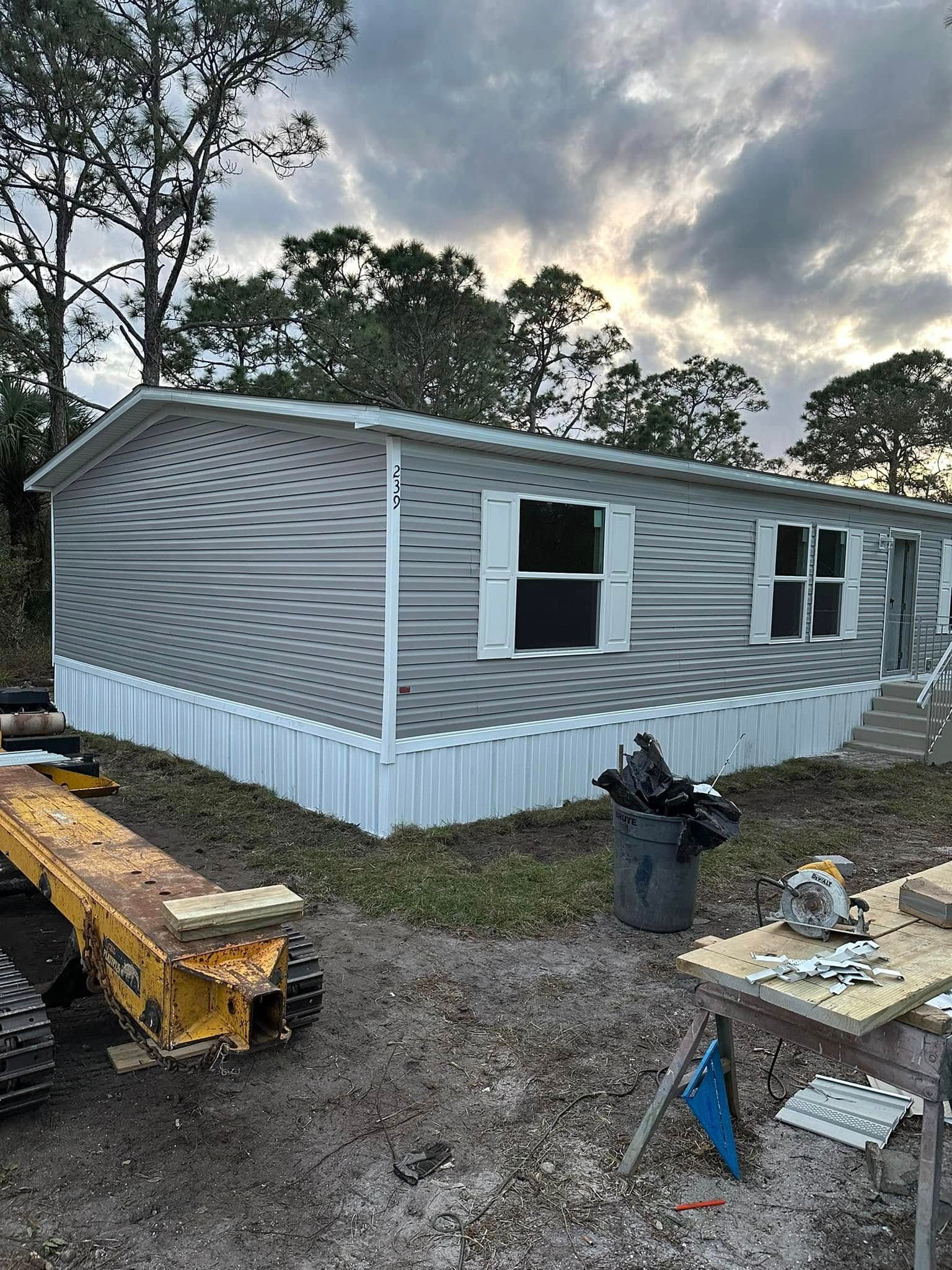 A gray mobile home with a wavy pattern on its siding stands on a dirt lot with construction equipment nearby.