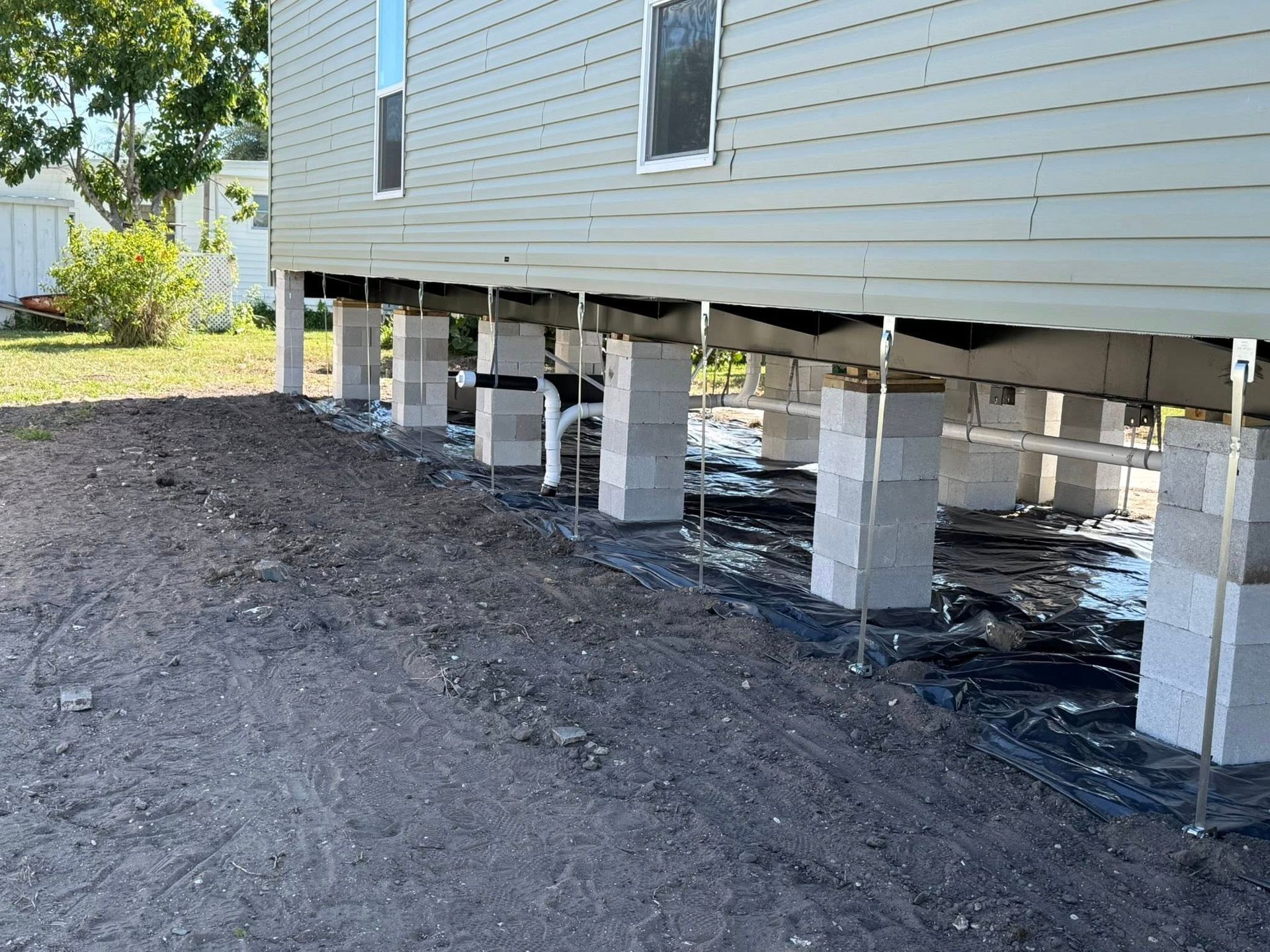 A side view of a house raised on cinder block pillars with a dark landscape fabric covering the ground underneath.