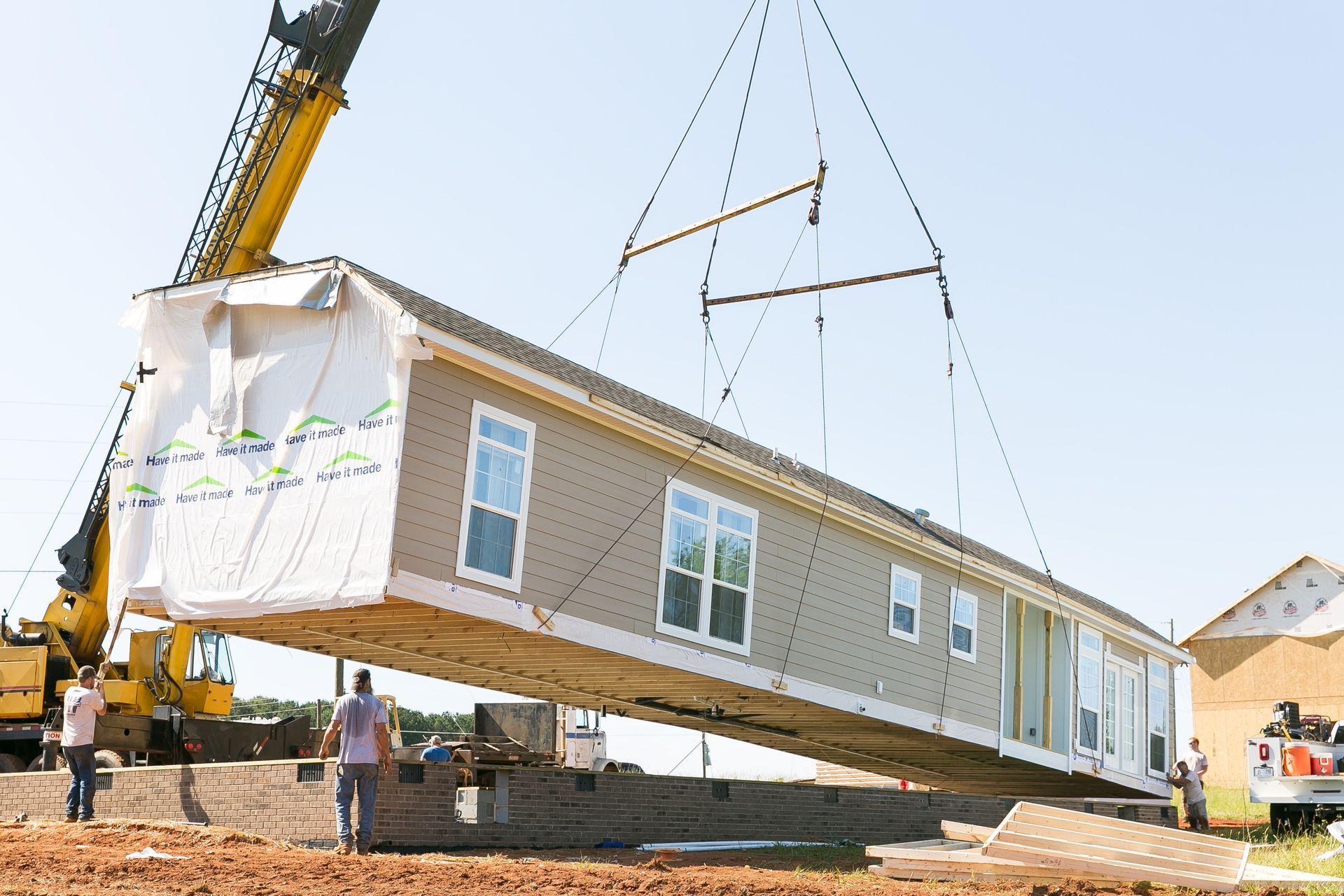 Crane lifting a beige modular house frame at a construction site under a clear sky