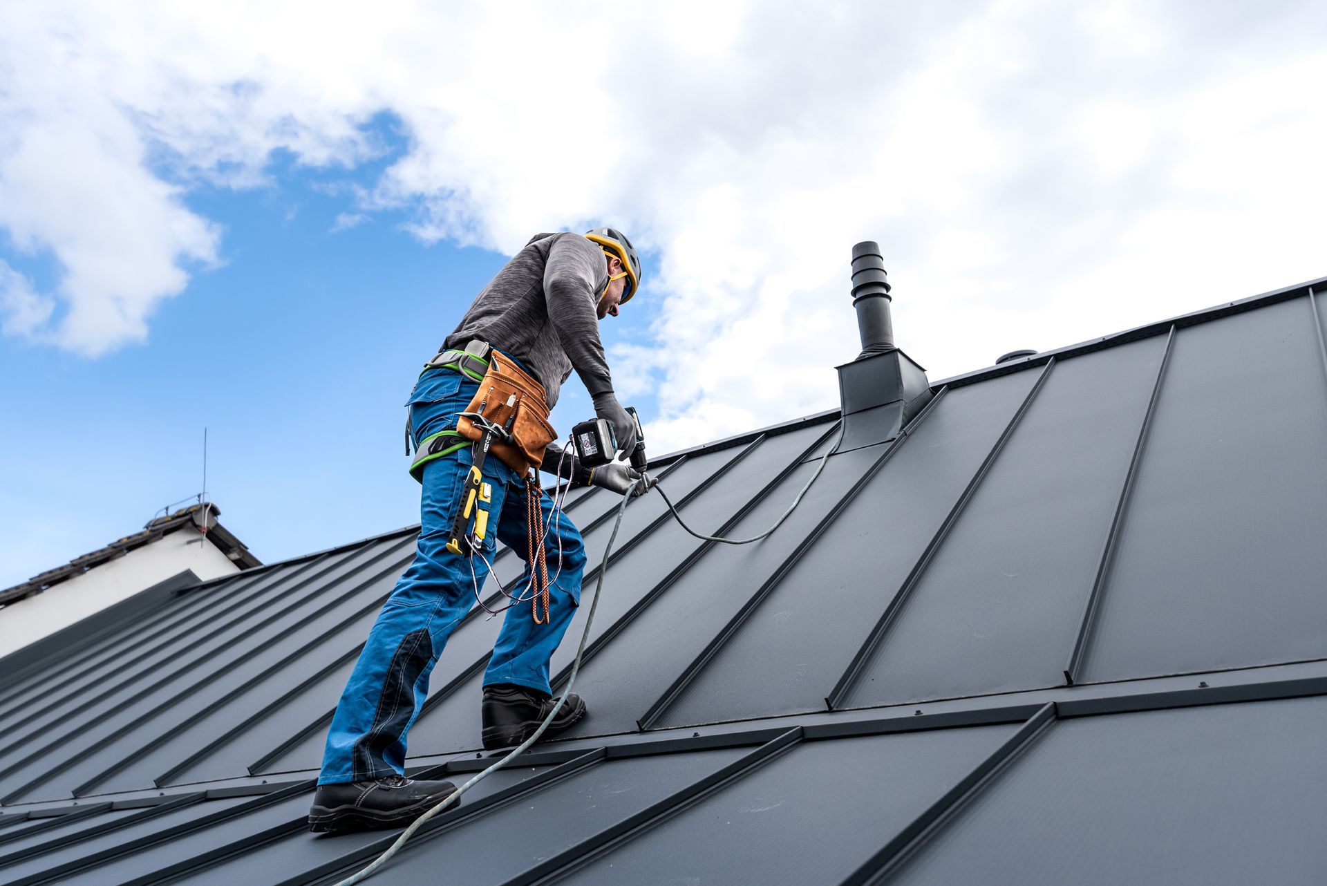 Roofer on a metal roof, installing something near a chimney, under a cloudy blue sky.