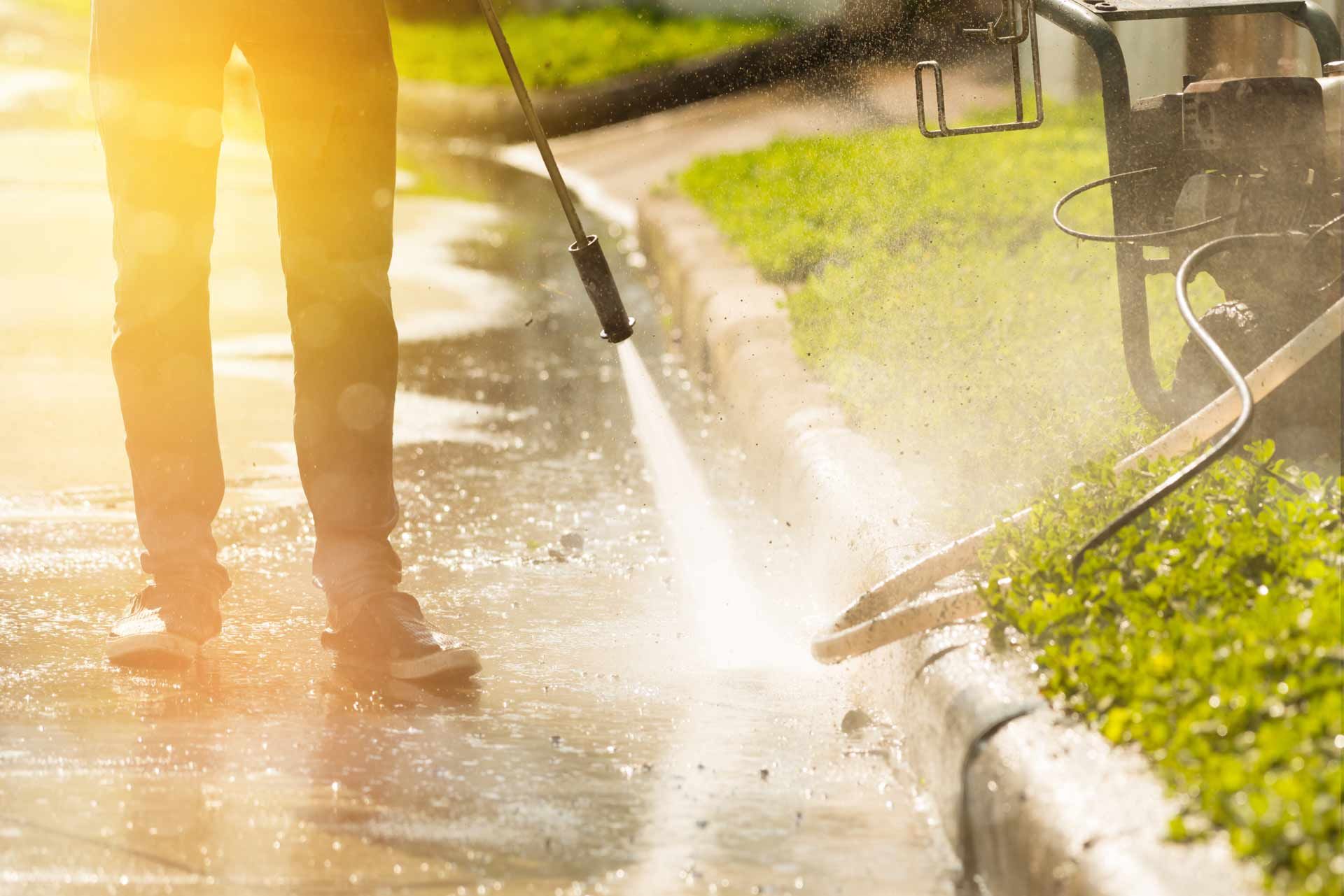 Person power-washing a sunlit sidewalk beside grass and a curb