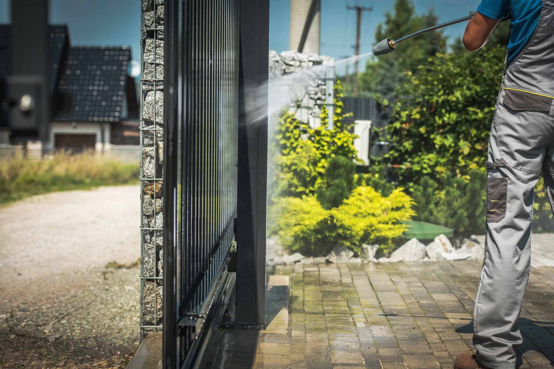 Person pressure-washing a stone walkway beside a black gate and green garden.