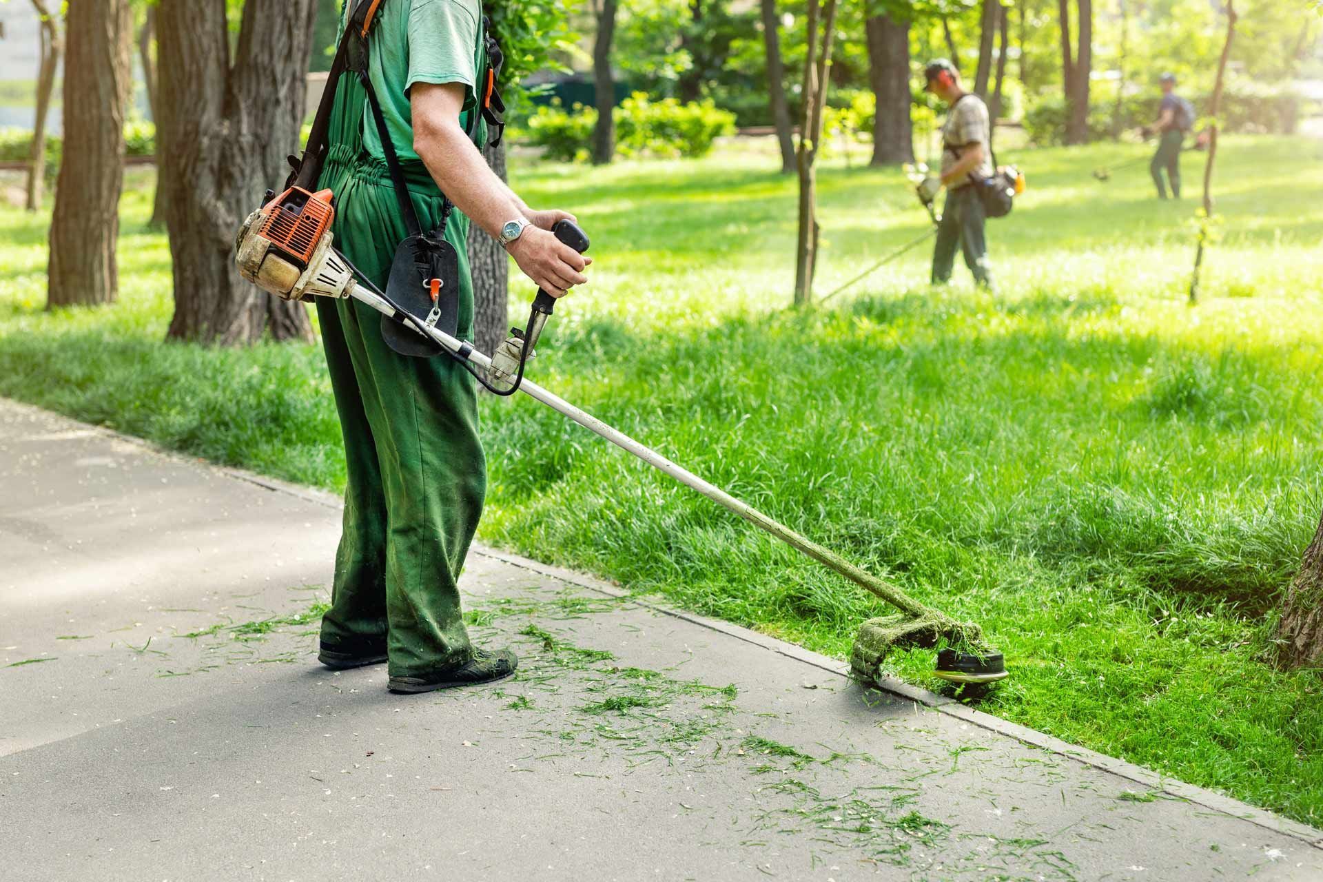 Person using a string trimmer to edge a grassy path in a park
