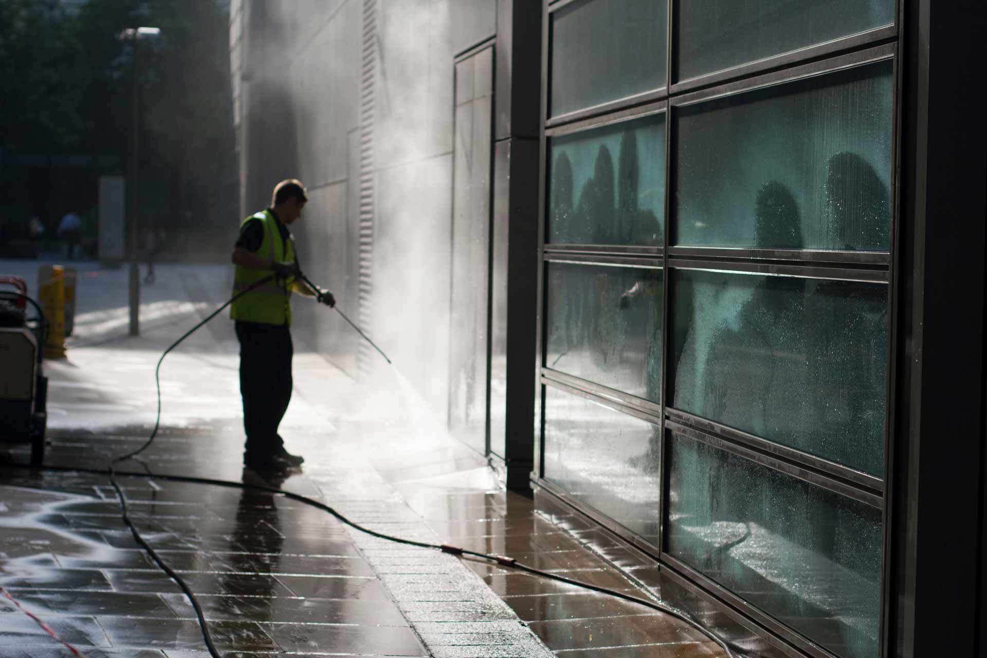 Worker pressure-washing a glass building exterior beside a wet sidewalk in bright sunlight