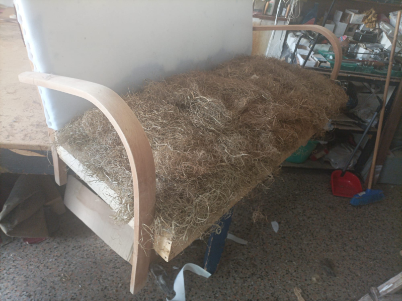 Una sedia con braccioli aperti, struttura in legno chiaro, schienale bianco e seduta imbottita con materiale fibroso marrone, in un ambiente da laboratorio.