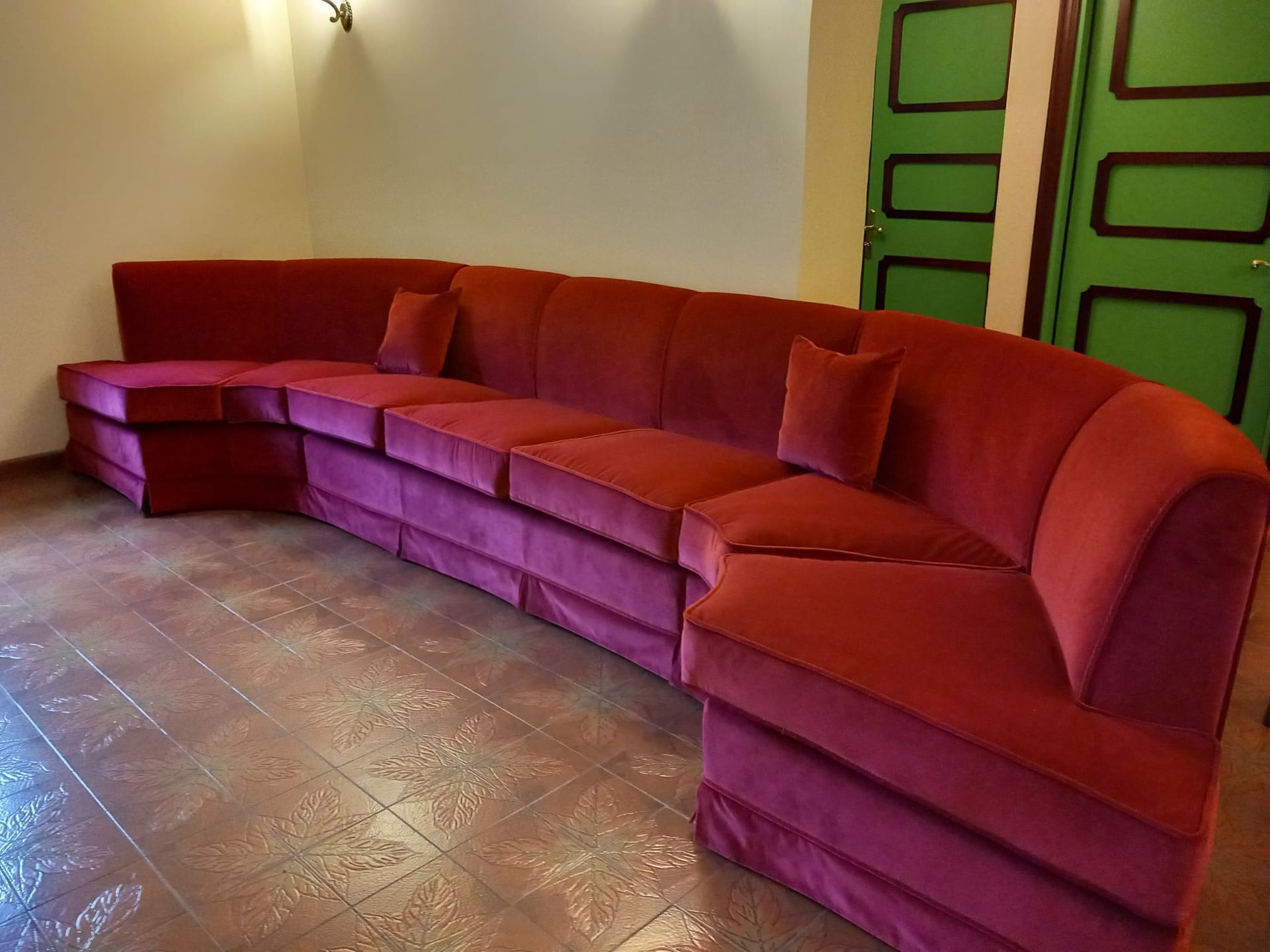Un grande divano curvo in velluto rosso in una stanza con porte verdi e pavimento in piastrelle marroni.