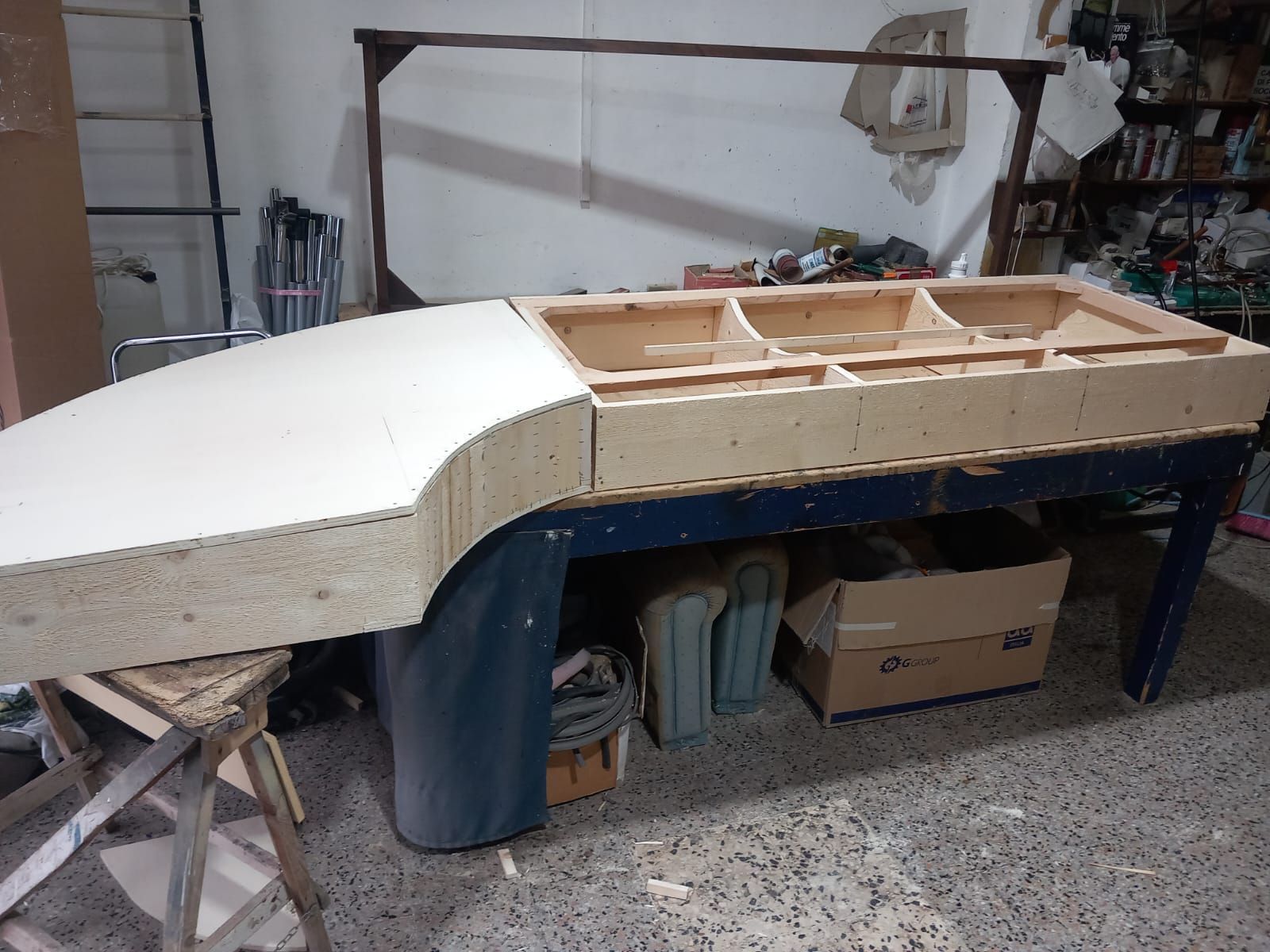 Una scrivania o un piano di lavoro in legno parzialmente costruito. È realizzato in legno chiaro su un tavolo con gambe blu, in un ambiente da laboratorio.