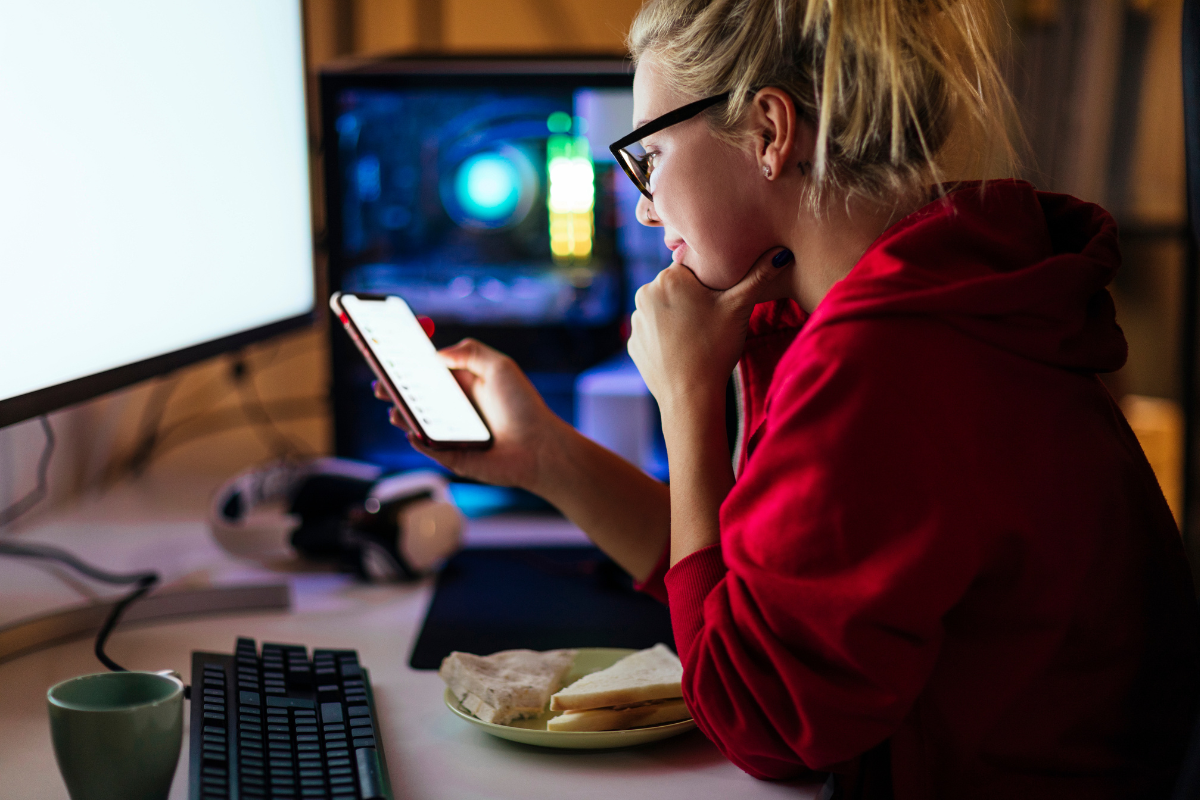 A woman in glasses and a red hoodie checks her smartphone while seated at a desk with a computer, keyboard, and sandwich.