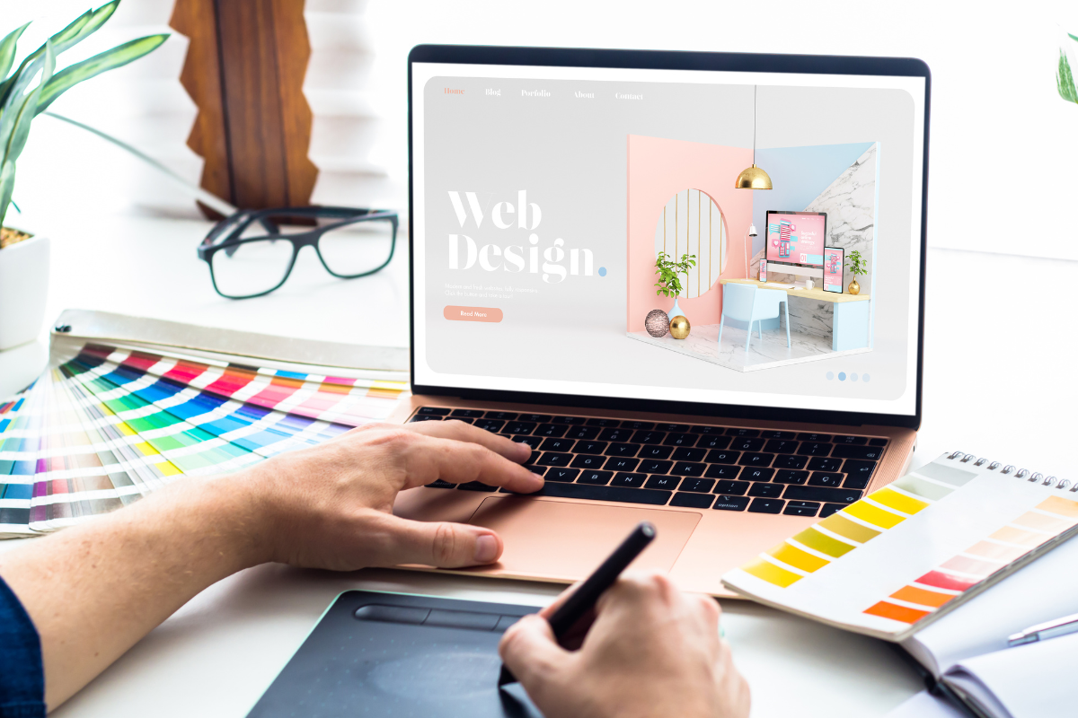 Designer working on a web design project on a laptop, surrounded by color swatches and using a graphics tablet.