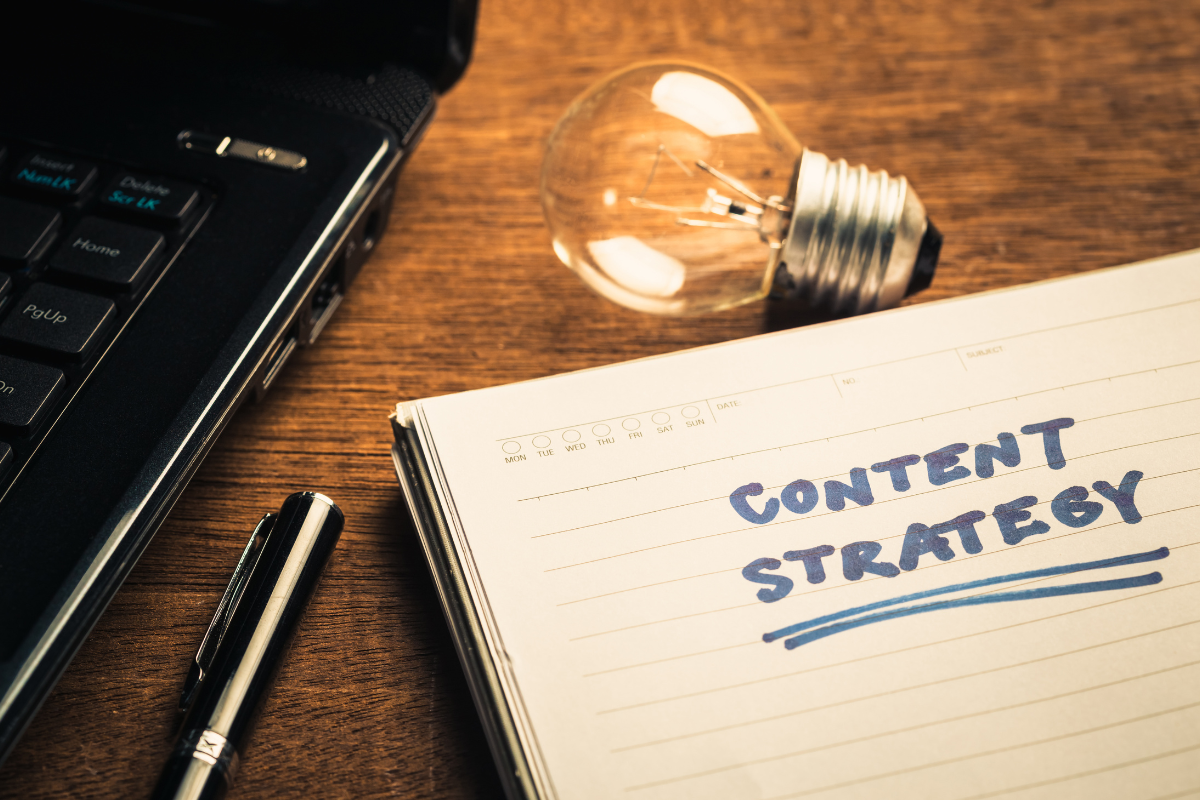 A laptop, pen, and lightbulb placed next to a notebook with the words 'Content Strategy' written on the first page.