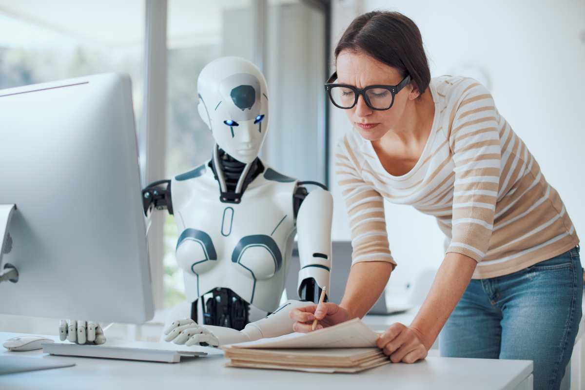 A woman working with a humanoid robot at a computer, reviewing documents while the robot types on a keyboard.
