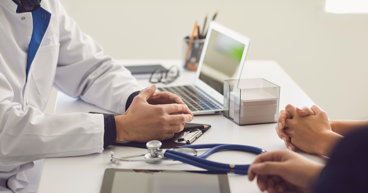 A doctor consulting a patient at a desk with a stethoscope, tablet, and laptop, representing healthcare services.