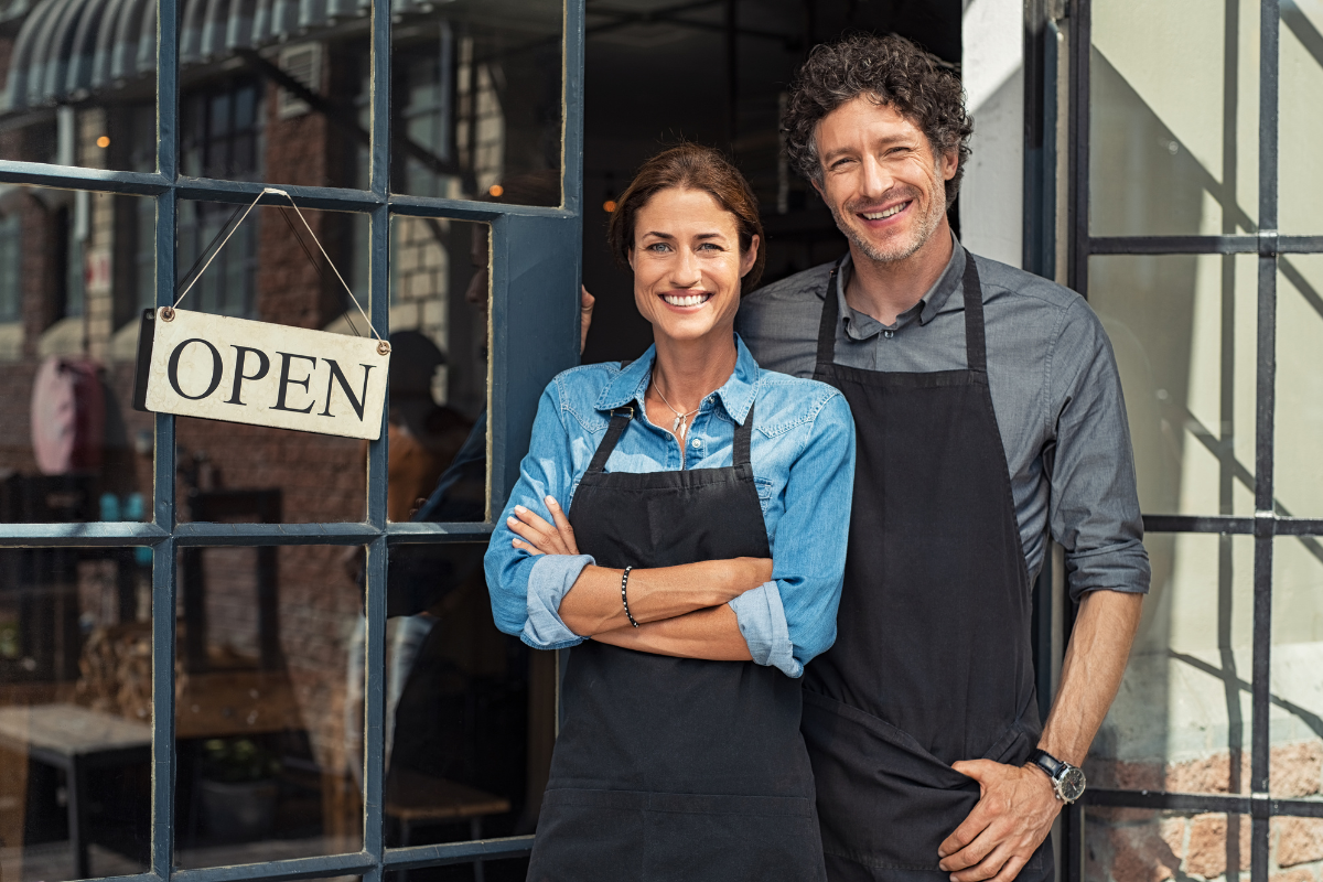 A smiling small business owner couple in aprons standing outside their store with an 