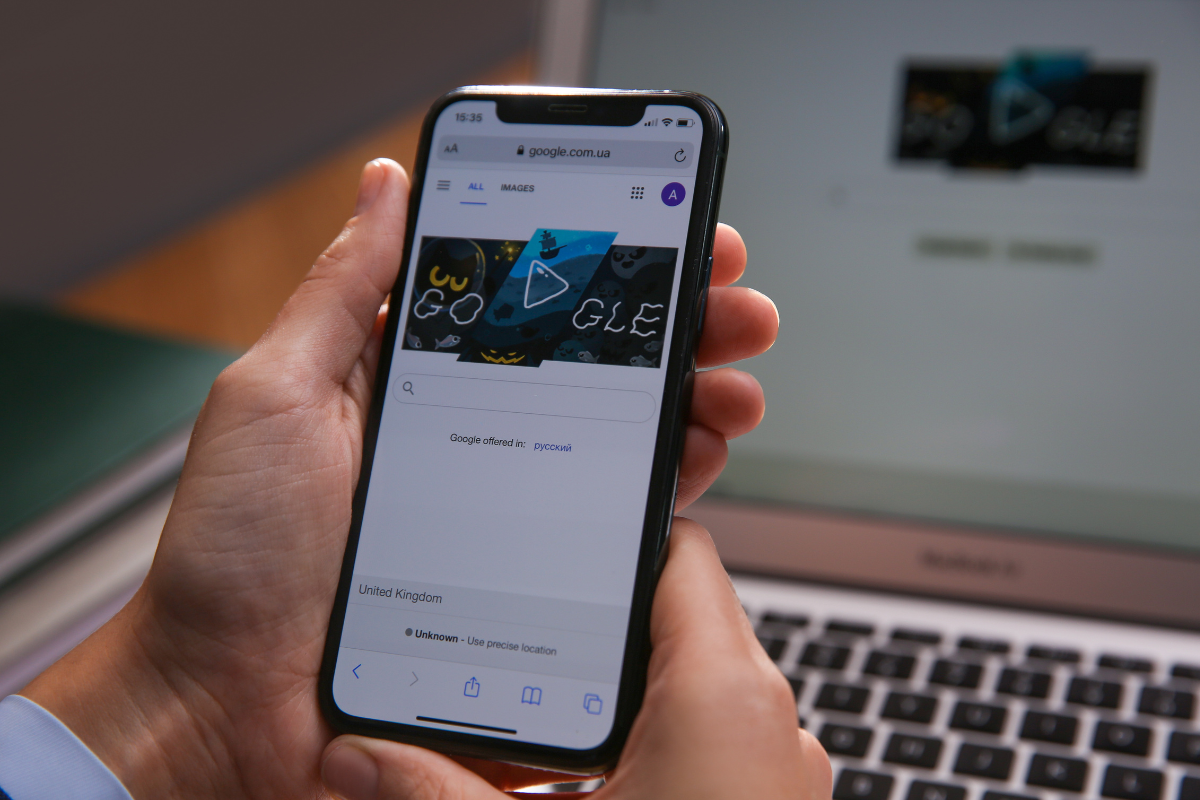 A person holding a smartphone displaying the Google homepage, with a laptop showing the same screen in the background.