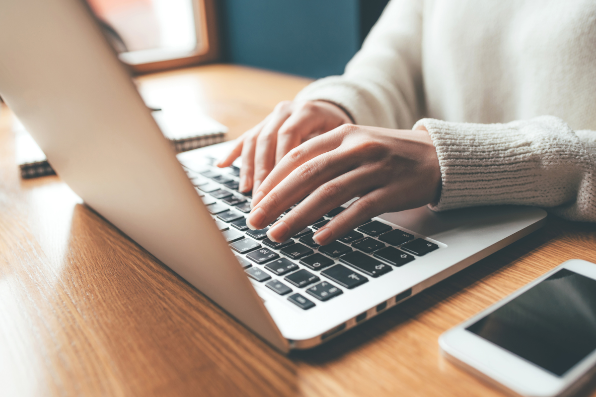 A person typing on a laptop at a wooden desk, with a smartphone nearby, symbolizing digital work or remote tasks.