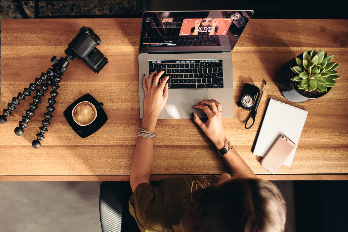 A content creator edits a vlog on a laptop, surrounded by a camera, tripod, notebook, and coffee on a wooden desk.