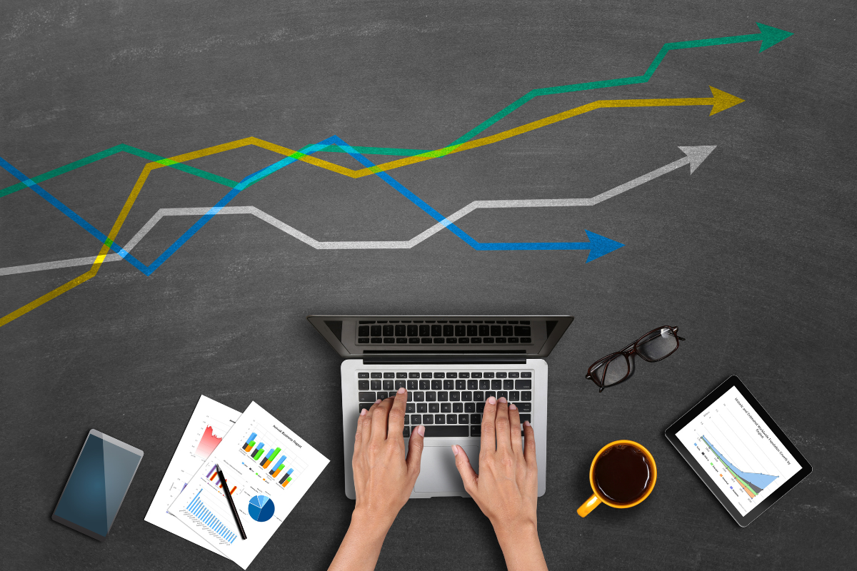 Hands typing on a laptop surrounded by charts, graphs, a smartphone, a tablet, and a coffee cup, with upward arrows above.