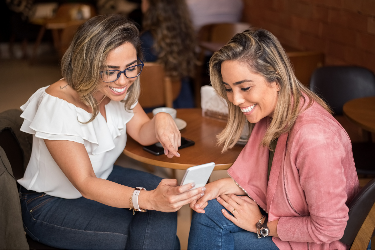 Two smiling women sitting at a café, looking at a smartphone and sharing content while enjoying their conversation.