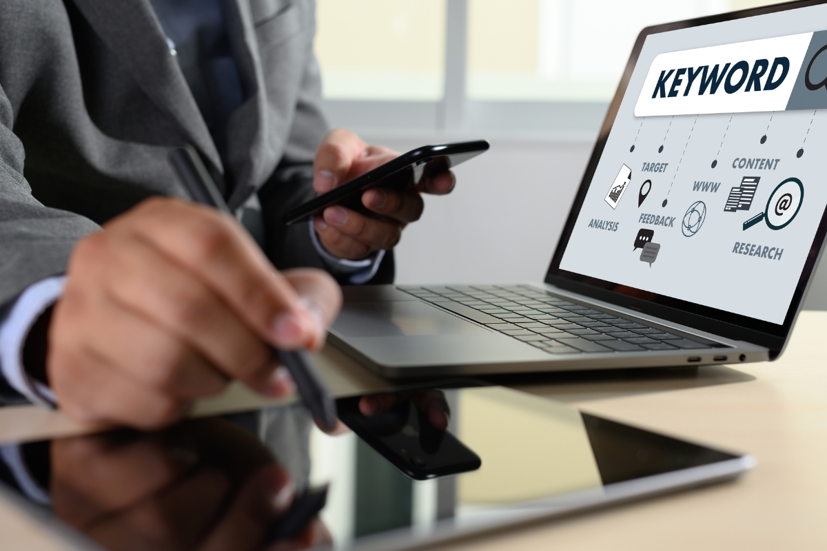 A person works with a laptop, phone, and tablet, analyzing keywords and SEO strategies displayed on the laptop screen.