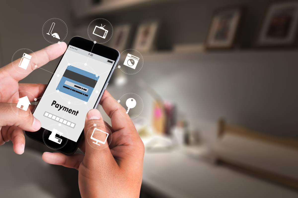 Hands holding a smartphone displaying a payment screen, surrounded by icons for smart home devices, representing digital transactions.