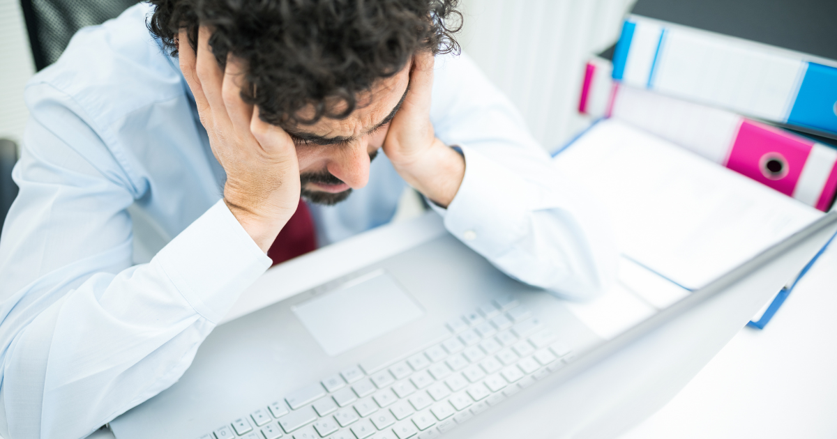 Stressed businessman holding head in hands at desk, frustrated while working on laptop in office setting.