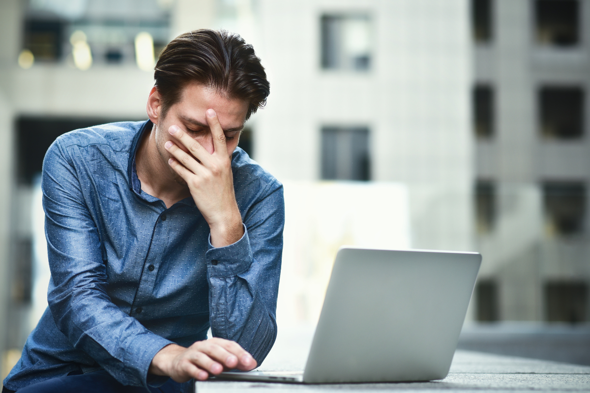 A frustrated man in a blue shirt sits outdoors with his hand on his face, looking at a laptop.