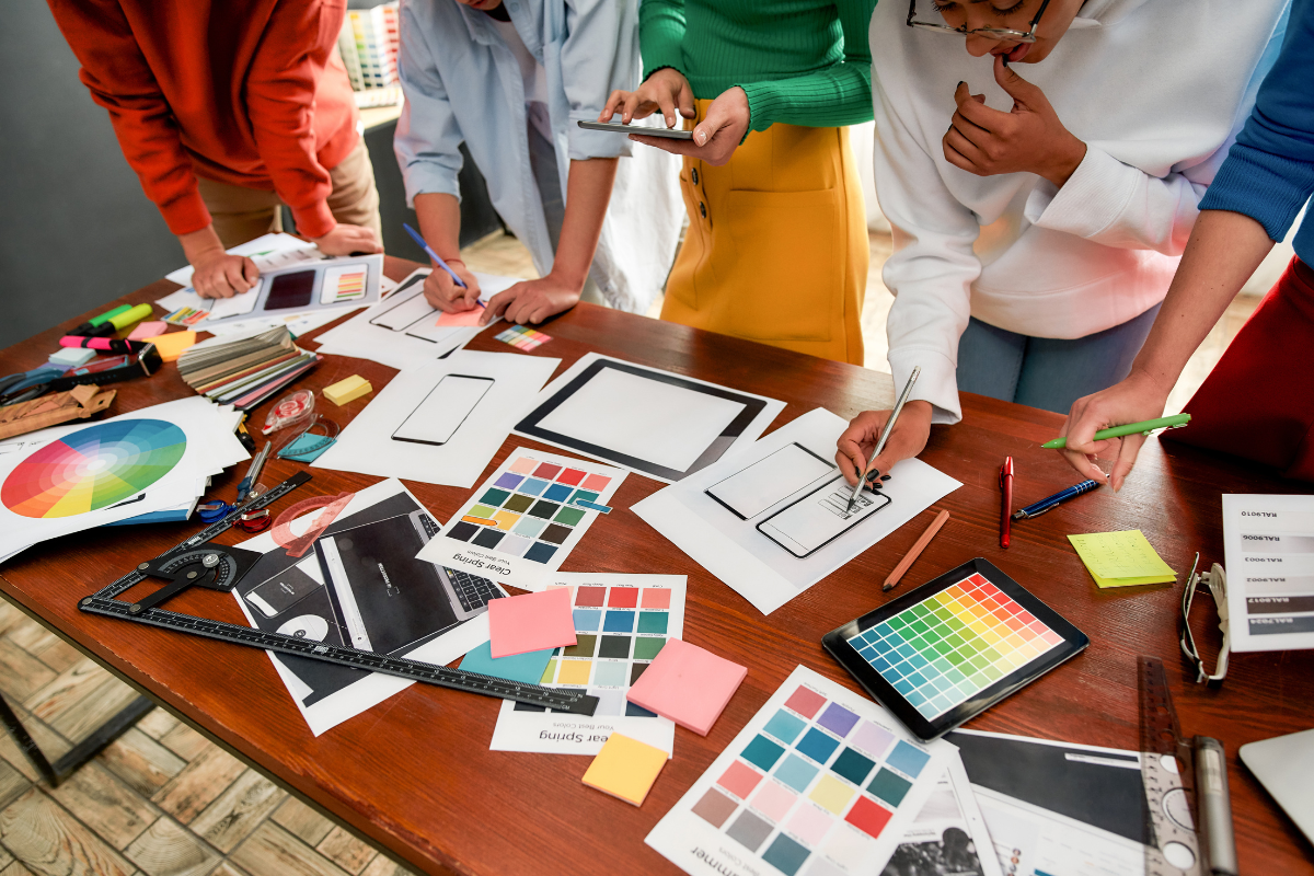 Design team collaborating at a table with color swatches, sketches, and digital tablets, planning web layouts and color schemes.