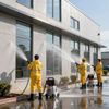Three workers in yellow suits pressure washing a building's exterior with silver machines.