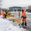 Workers in orange suits clear snow from a parking lot with a yellow tractor and shovels.