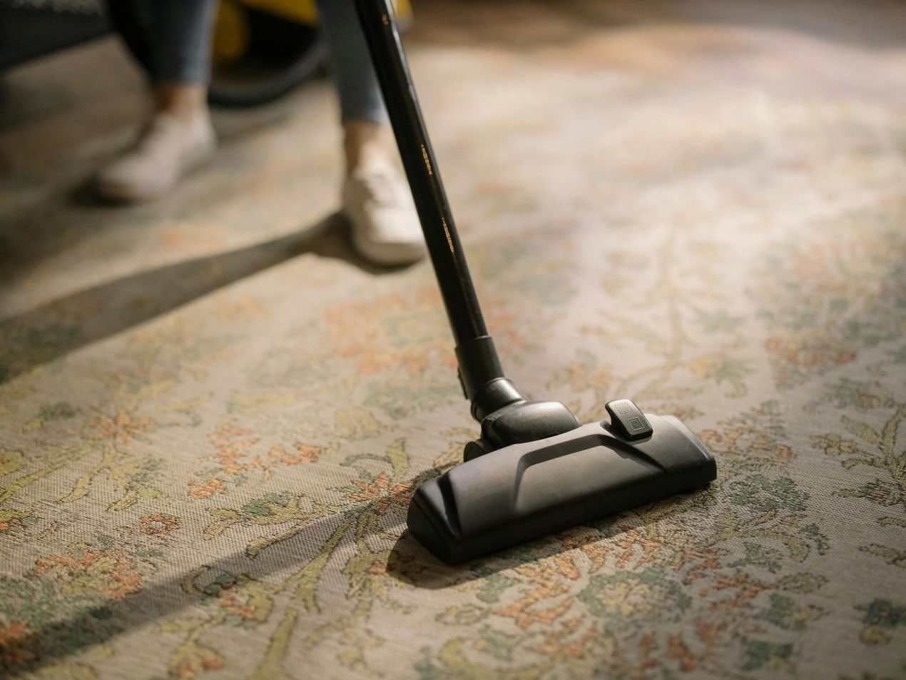 A person using a vacuum cleaner to clean a patterned carpet in a softly lit room.