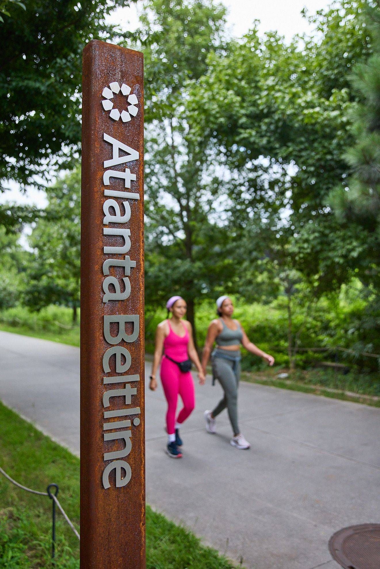 Two walkers on a park path beside an Atlanta Beltline sign surrounded by trees