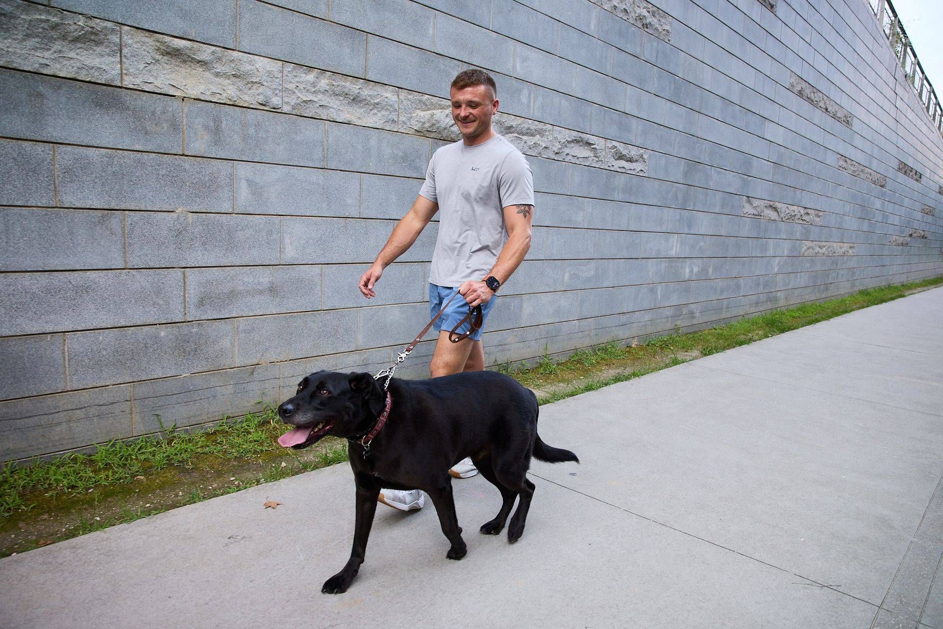 Man walking a black dog on a leash along a sidewalk beside a large stone wall.