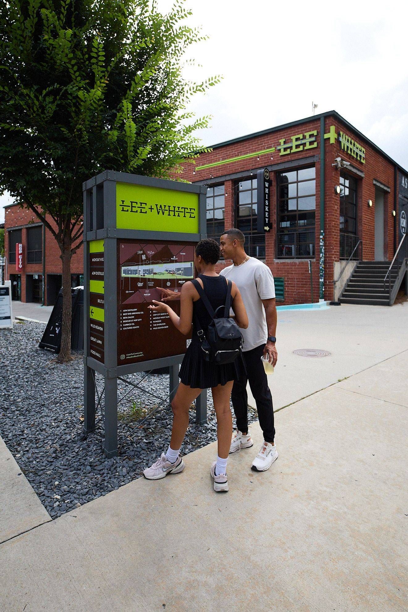 Three people read a sign outside a brick building with outdoor stairs and trees nearby.