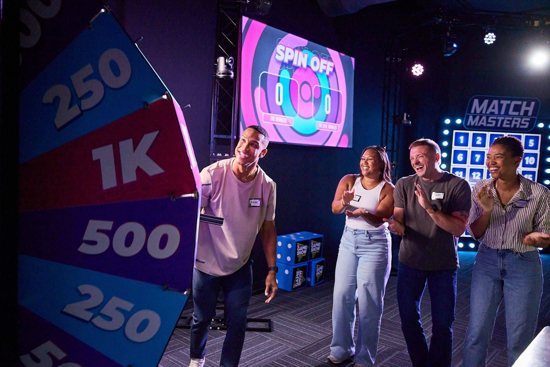 People on a game show stage beside a colorful prize wheel and illuminated screens
