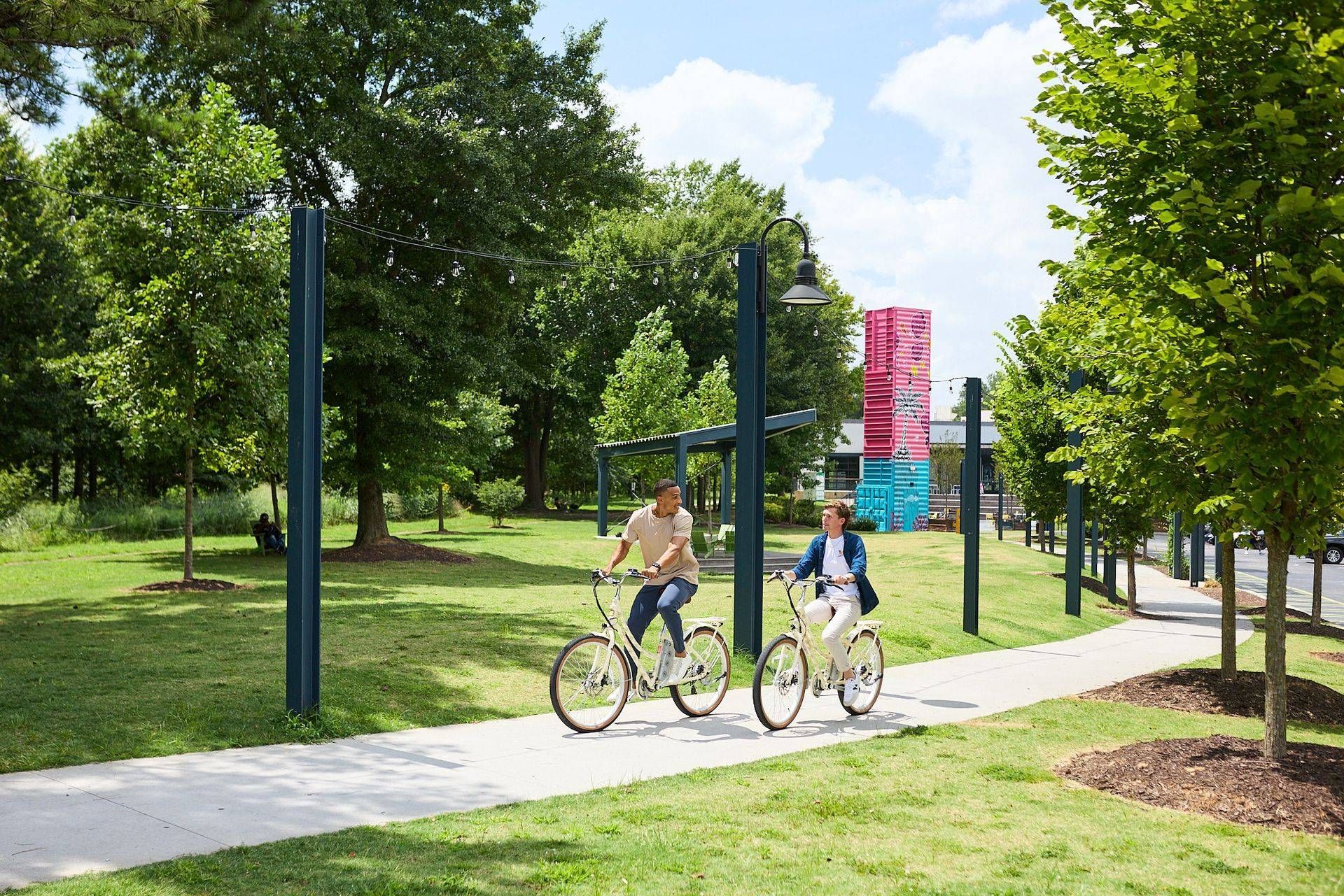 Two cyclists ride on a path through a green park on a sunny day.