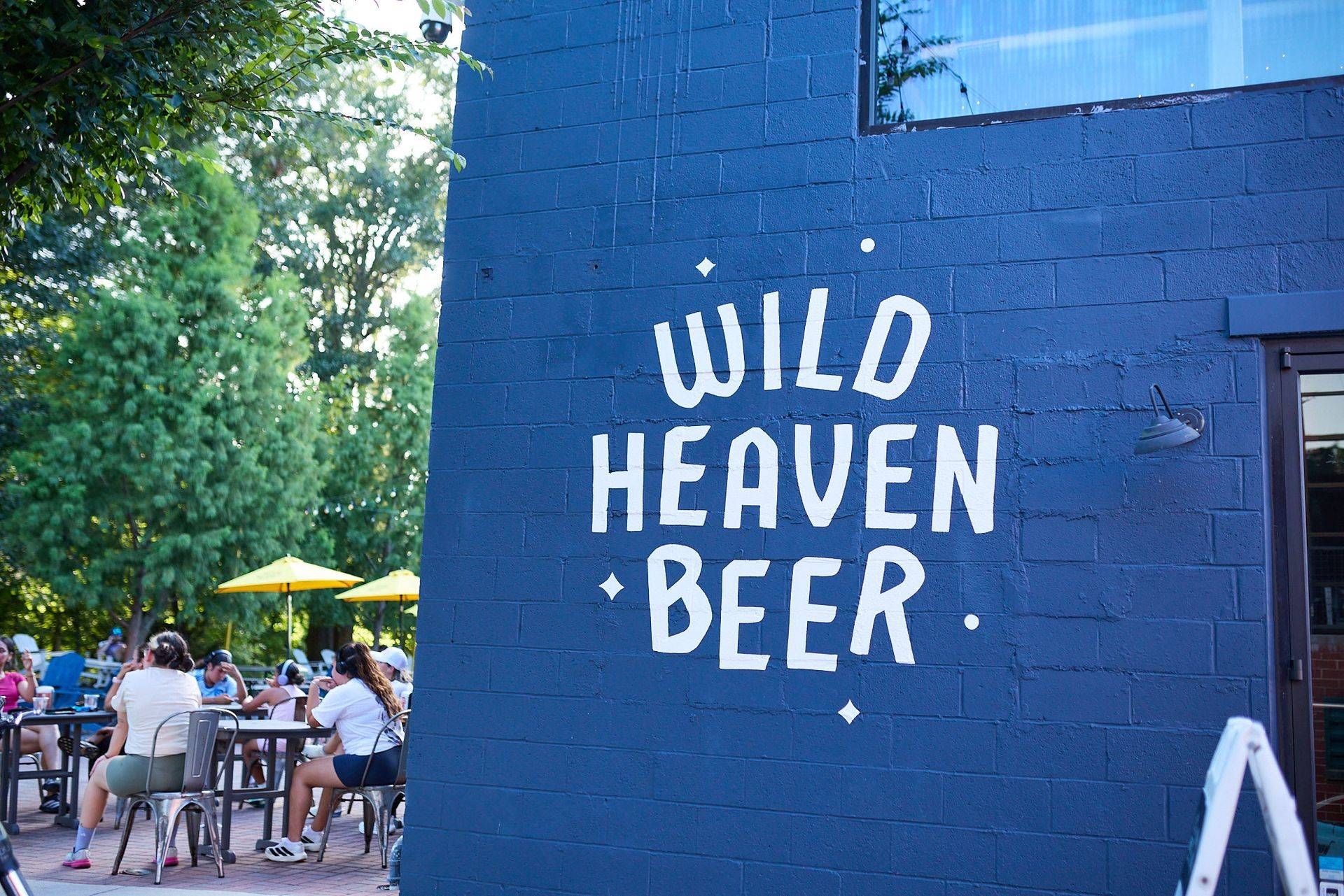 Blue wall with “WILD HEAVEN BEER” painted on it beside a sunny outdoor seating area