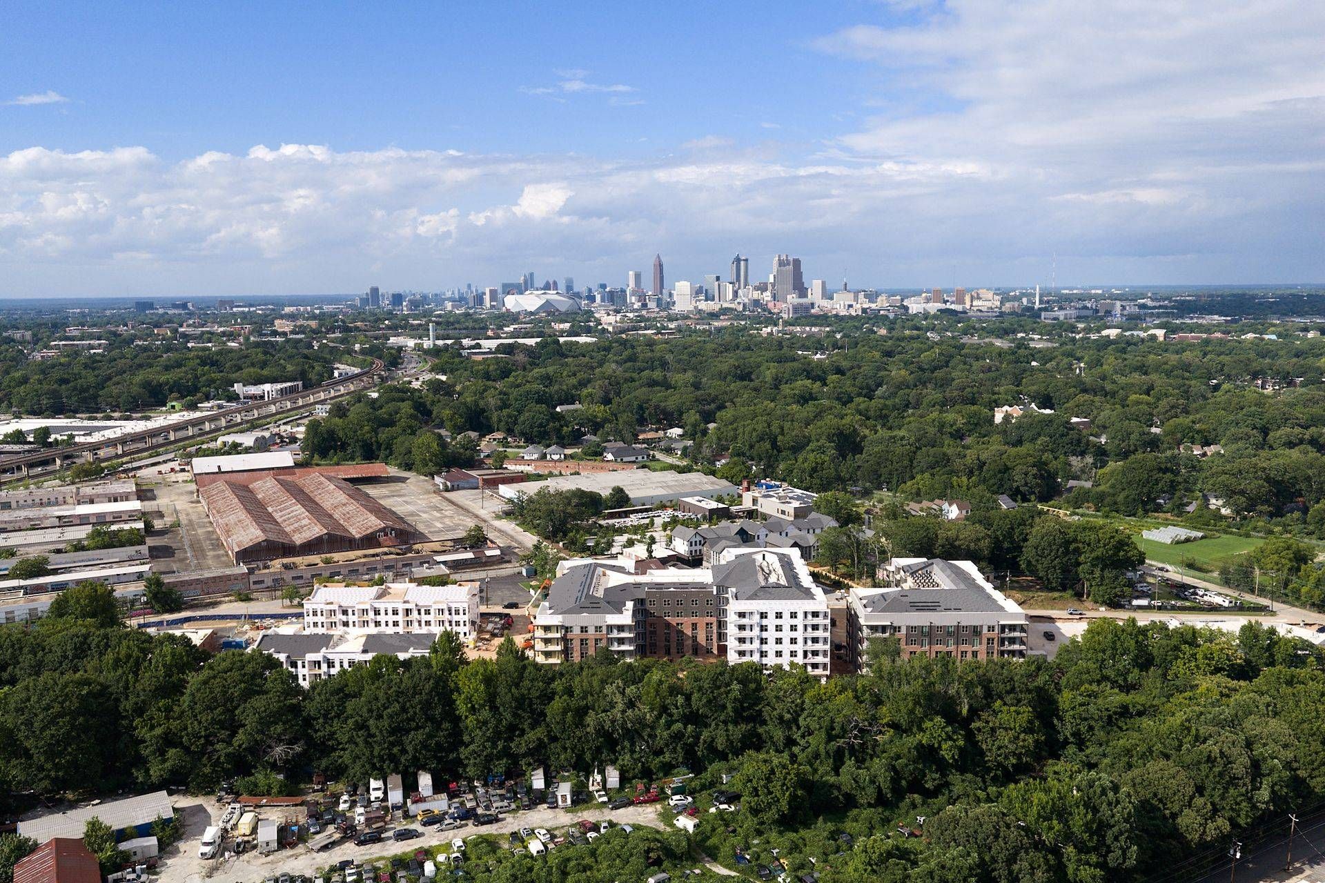 City skyline beyond a green forest and buildings under a partly cloudy blue sky