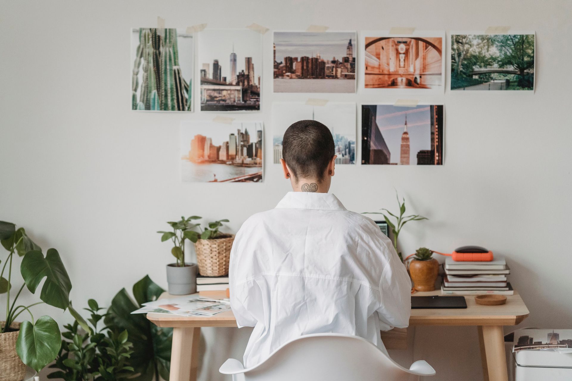Person working at a desk with photos pinned on a wall, surrounded by plants and a laptop